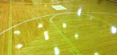 Indoor gymnasium with polished wooden basketball court floor, marked with green and white game lines under bright overhead lights.