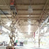 Worker on a scissor lift cleaning a high industrial ceiling with bright overhead lights and exposed beams.