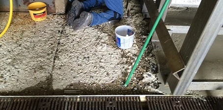 Person in work clothes and boots lying partially hidden on a rough concrete floor near a bucket, hose, and tools, suggesting a hazardous or unsafe work situation.
