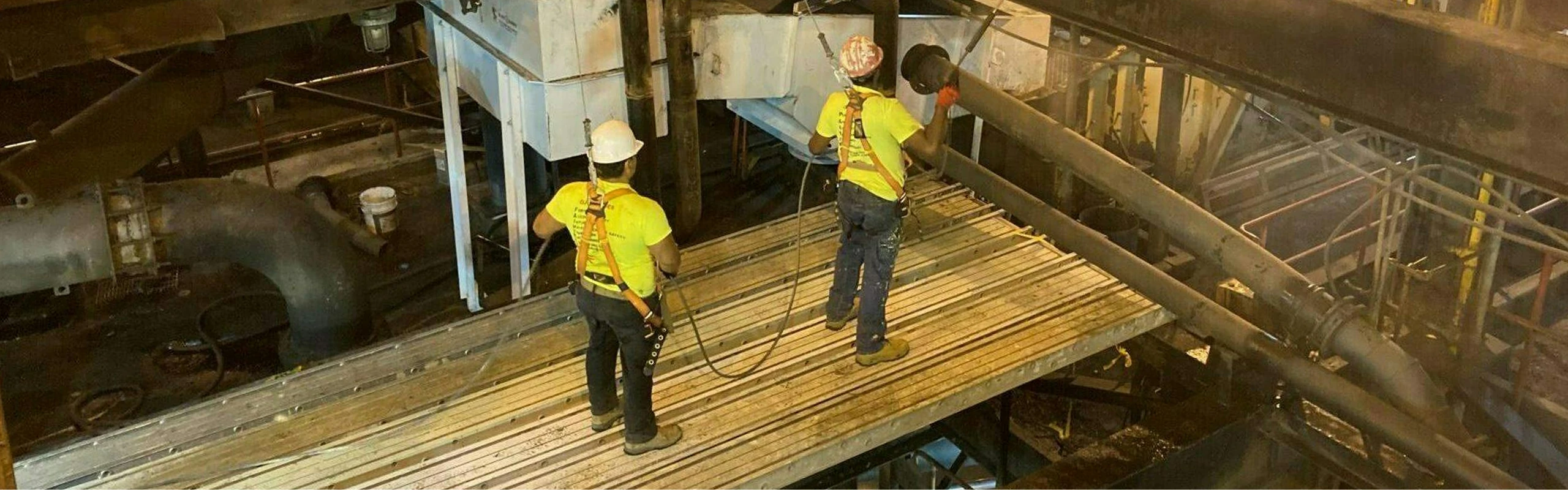 Two workers wearing safety harnesses and bright yellow shirts stand on a grated platform inside an industrial facility, inspecting large metal pipes and machinery under warm lighting.