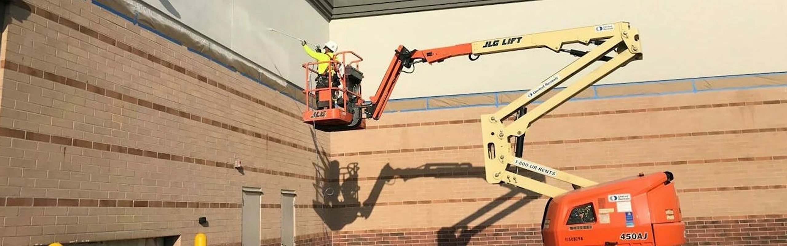 Worker in safety gear on a boom lift painting the upper wall of a brick and concrete commercial building.