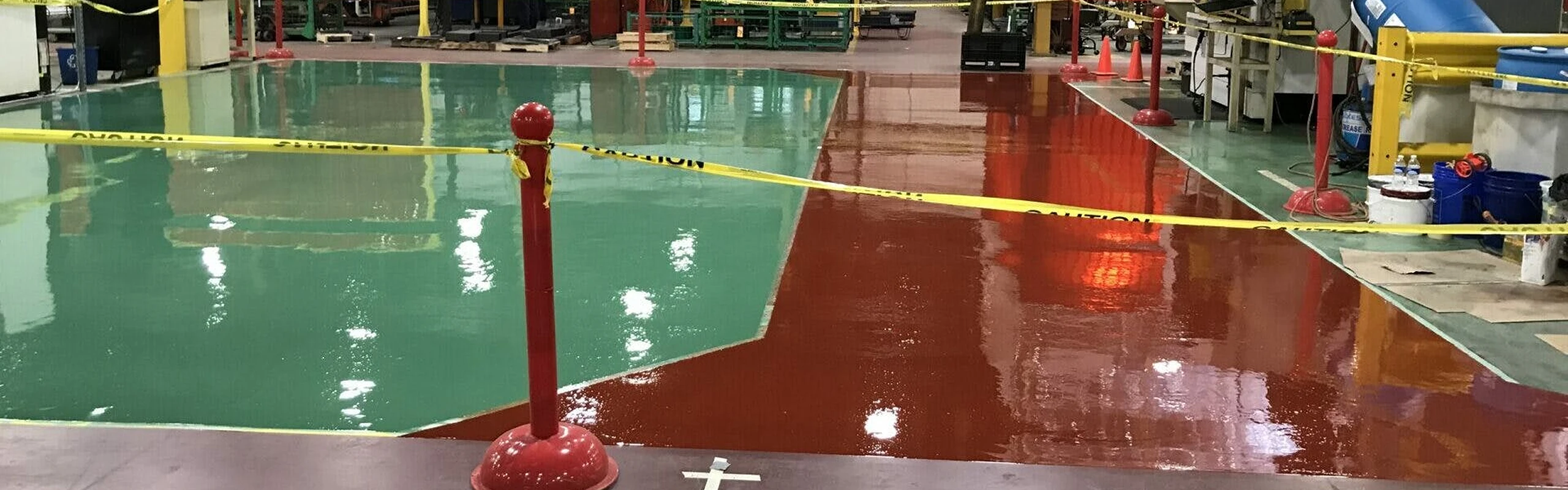 Fresh green and red epoxy floor coating curing in a production area, roped off with yellow caution tape and red stanchions.