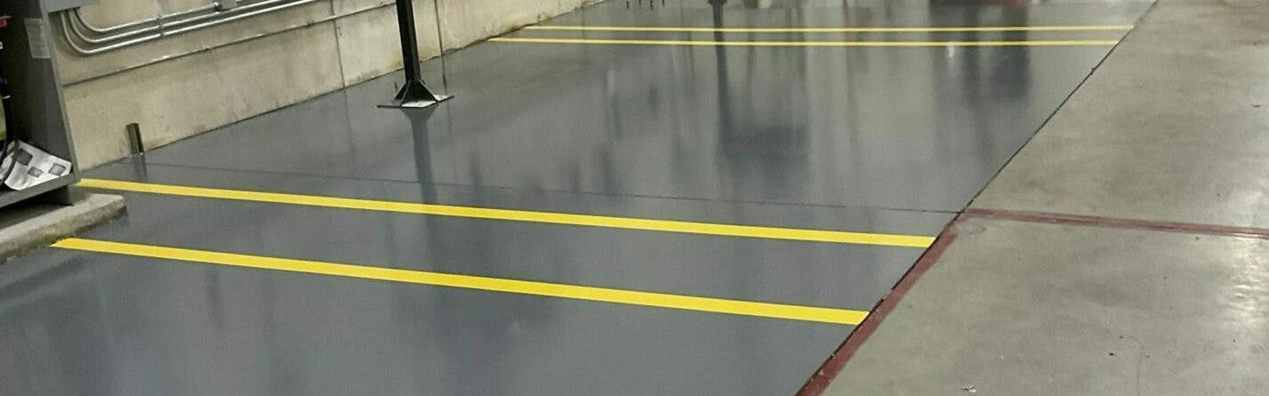 Smooth gray epoxy floor with bright yellow safety lane stripes beside machinery and conduit along a wall.