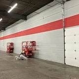 Worker painting a large warehouse interior wall with a bold red stripe, using lifts and equipment to reach high surfaces.
