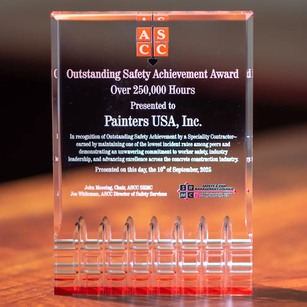 A clear rectangular acrylic award with red edges and a red base sits on a wooden surface. Near the top is the orange ASCC logo, followed by the engraved heading "Outstanding Safety Achievement Award Over 250,000 Hours," and the main text stating it is presented to Painters USA, Inc. for outstanding safety performance in the concrete construction industry, dated September 10, 2025, with small logos and signatory names along the bottom.