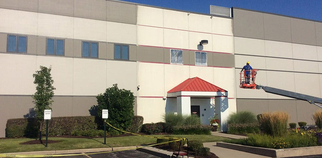 Worker on a boom lift paints the exterior of a beige commercial building with a red-roofed entry; windows are masked, caution tape lines the landscaped frontage, and the sky is clear.